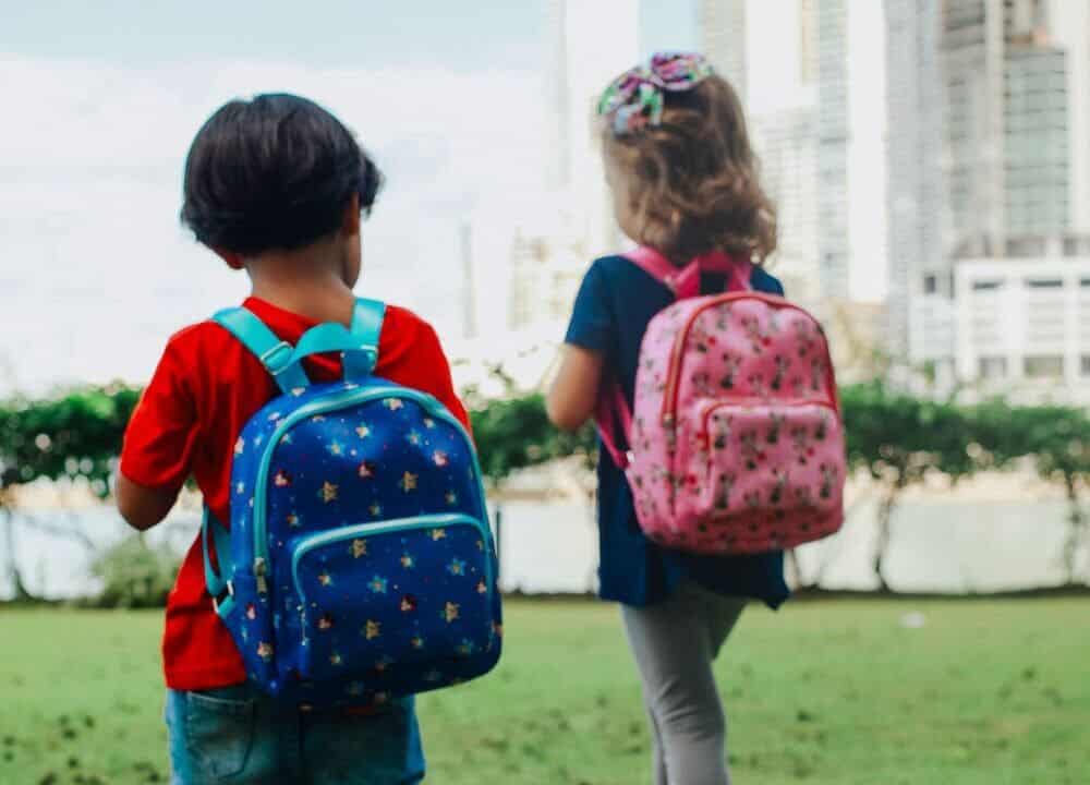 Two kids with colorful backpacks strolling through a city's green park in daylight.